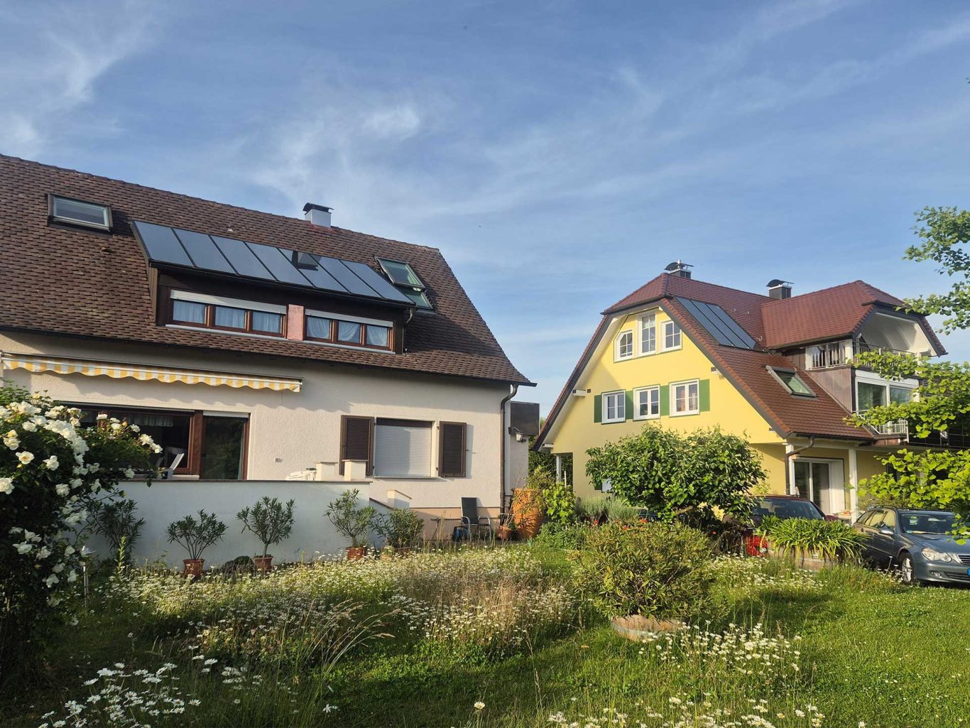 Ferienwohnungen Losch, Hagnau, Außenansicht Zwei Häuser mit grünen Gärten, blauer Himmel und Sonnenkollektoren auf dem Dach.